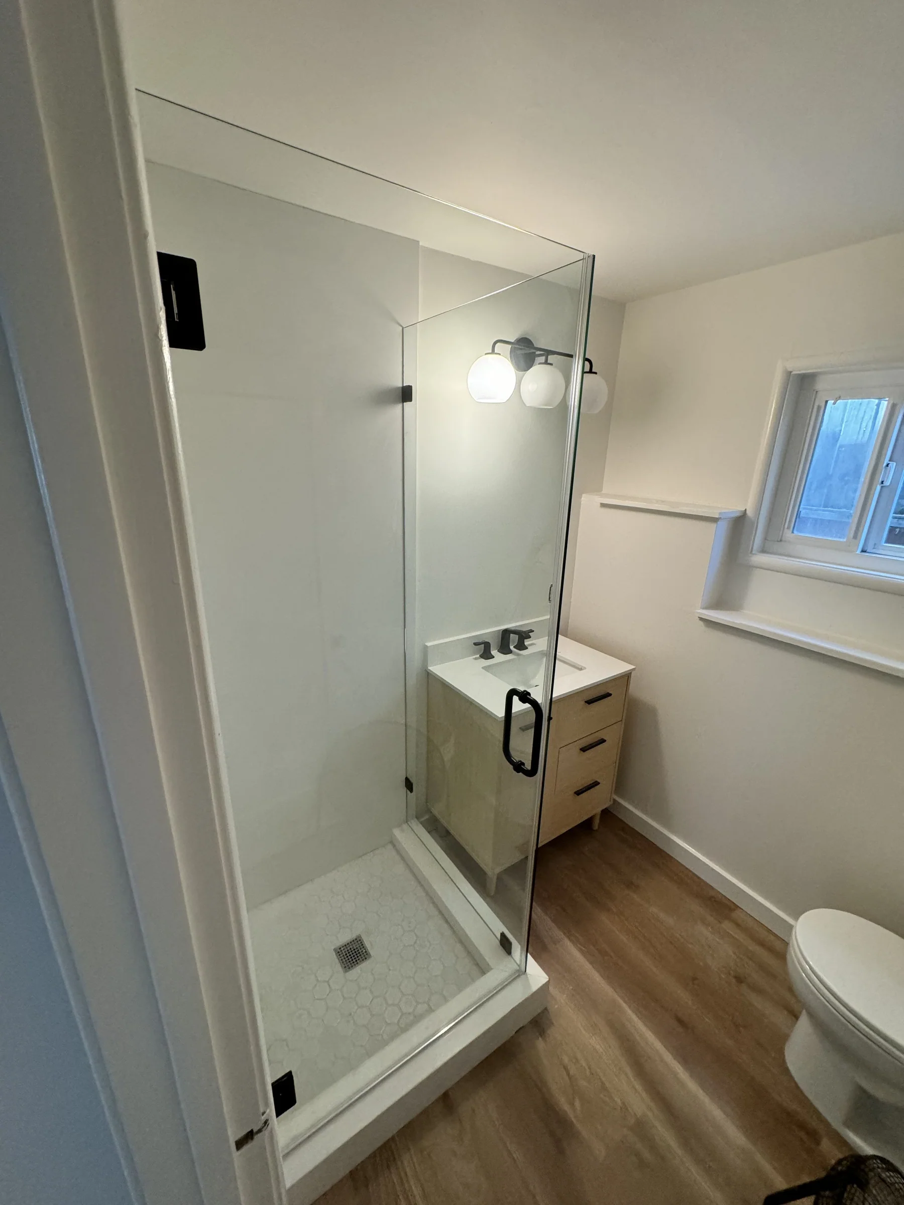 Bathroom remodel in Montclair with frameless glass shower enclosure, white hex tile shower floor, light wood vanity with black hardware, and three-globe sconce lighting