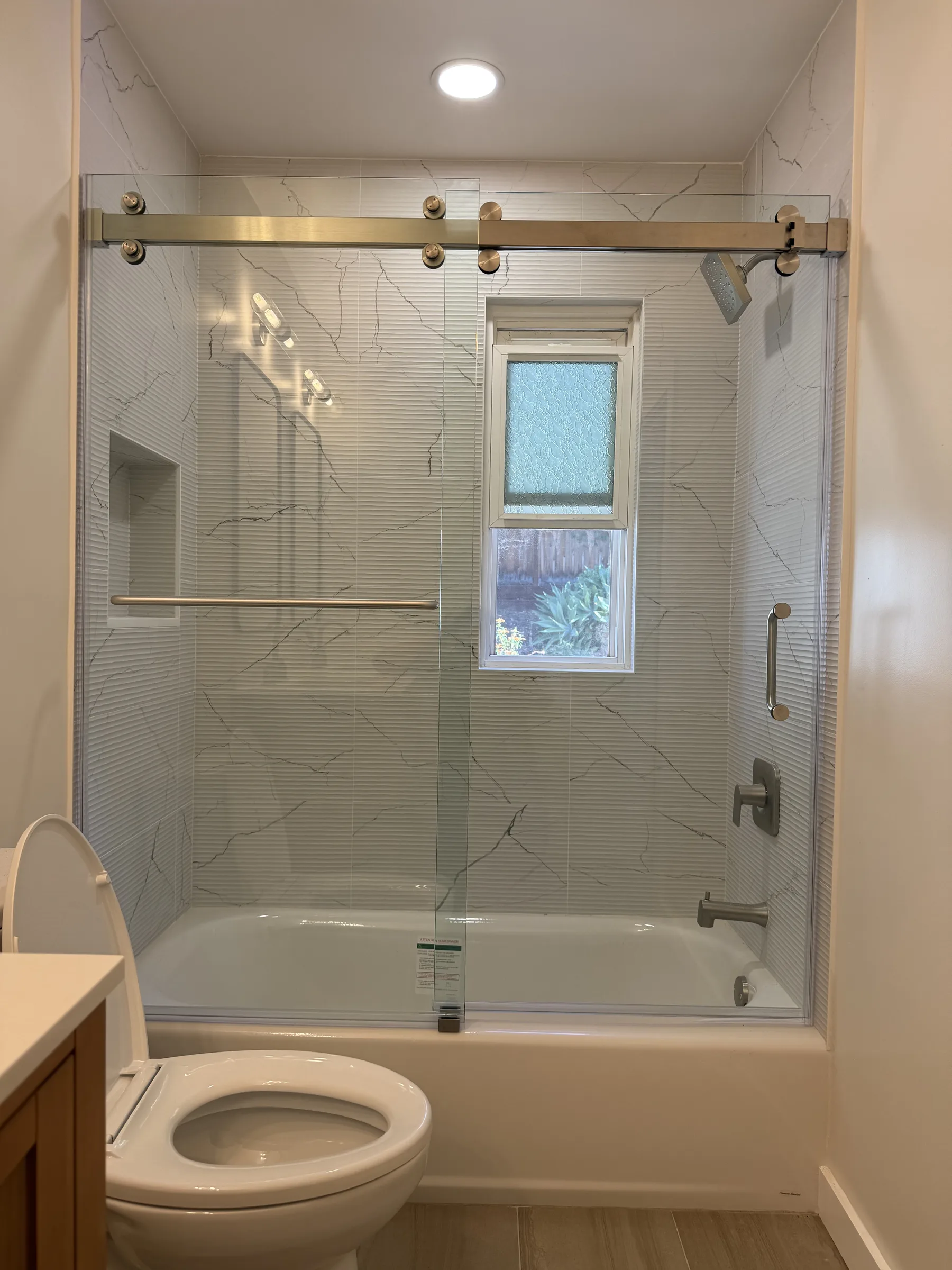 Finished Oakland bathroom tub with marble-look tile surround, brushed nickel sliding glass shower door, recessed niche, and window with privacy glass