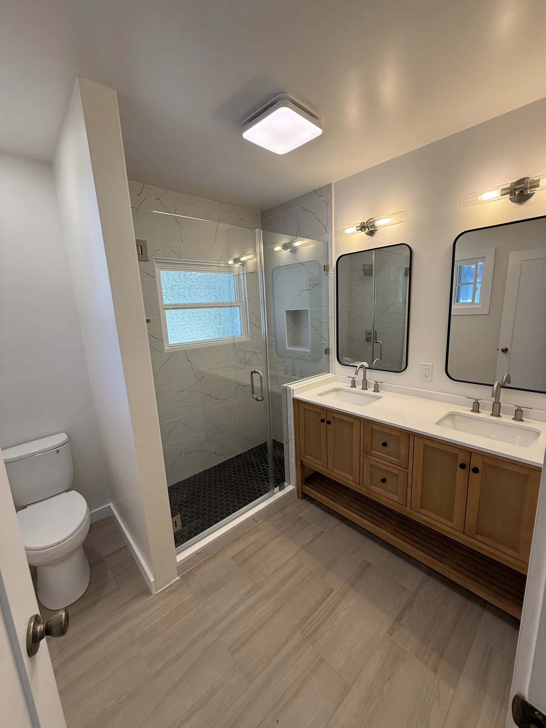 Finished Oakland bathroom with white oak double vanity, quartz countertop, black-framed mirrors, sconce lighting, and marble-look walk-in shower with black hex pan