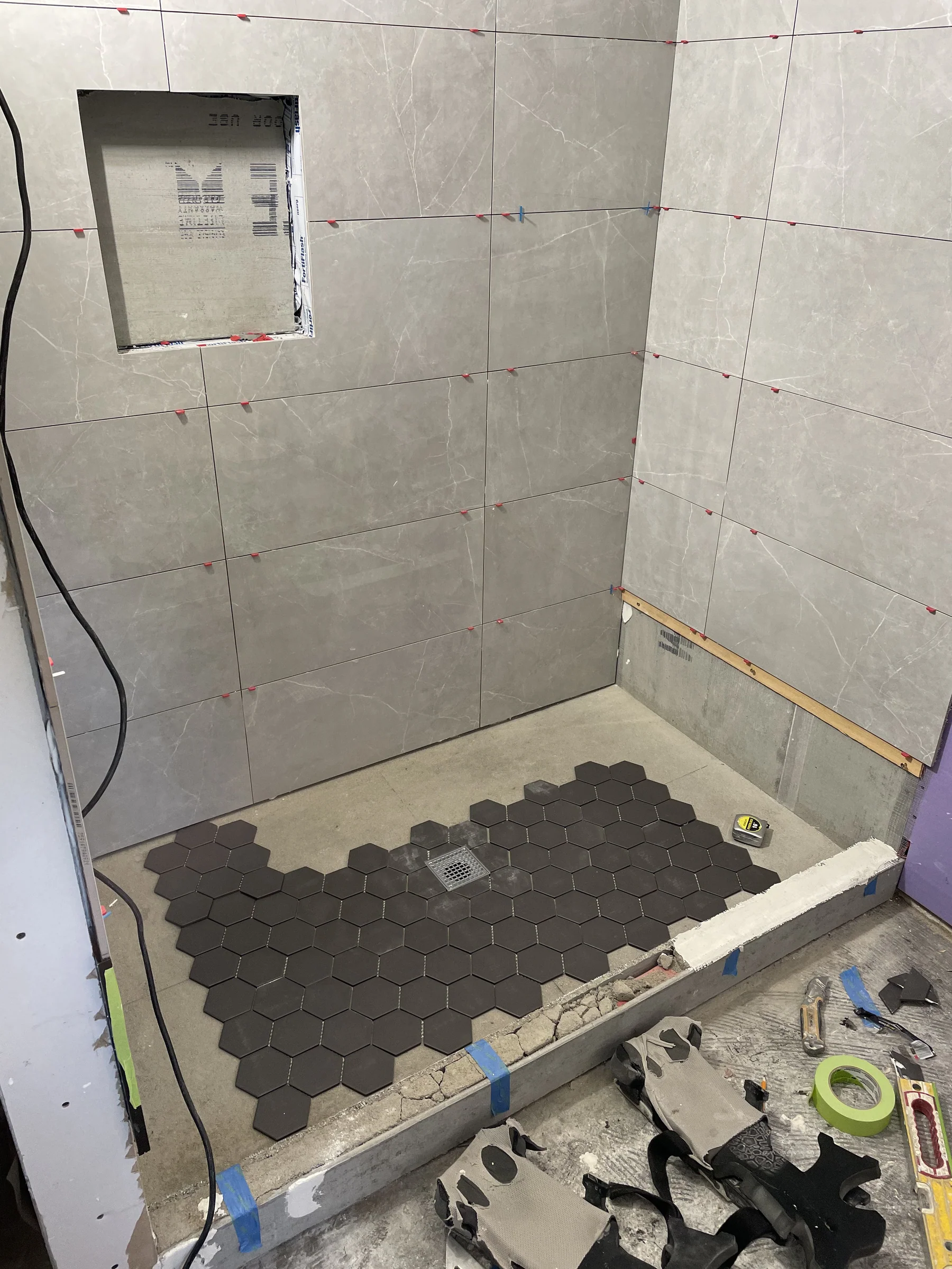 Bathroom shower tile installation in progress in a Berkeley remodel — gray marble-look wall tile with leveling clips, dark hexagon shower floor tile, and built-in niche framed out