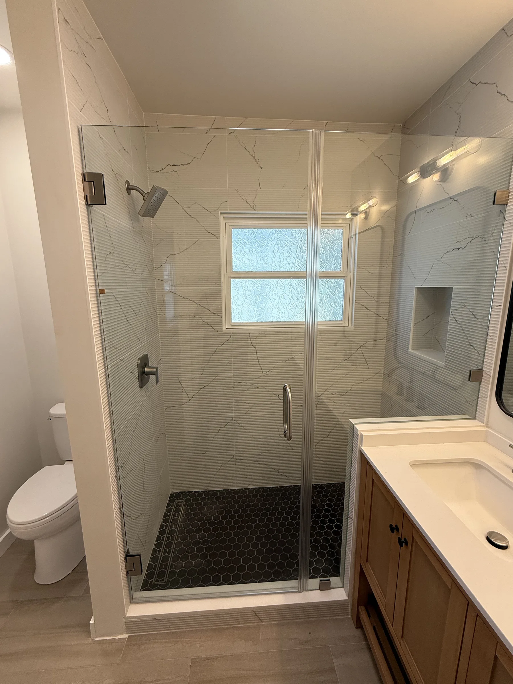 Finished Oakland walk-in shower with marble-look tile walls, black hexagon mosaic pan, frameless glass door, recessed niche, and quartz-top vanity