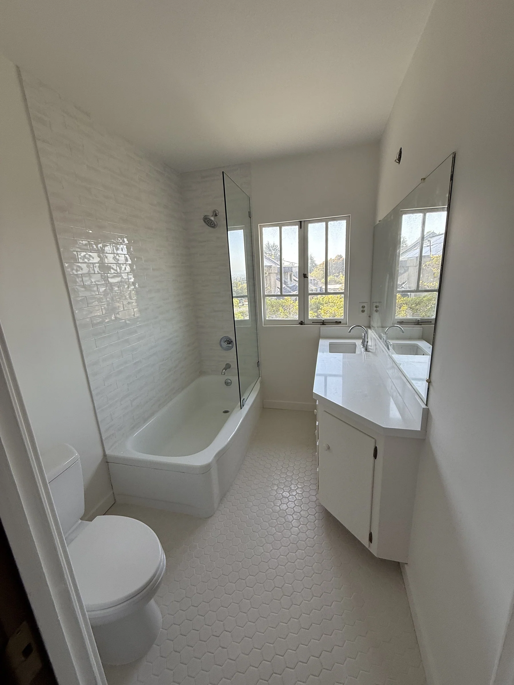 Finished Berkeley bathroom remodel — curved tub with half-height glass shower door, textured subway tile walls, white hex floor, double vanity, and Berkeley Hills view through casement windows