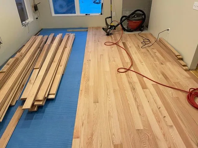 Hardwood floor installation in progress with oak planks, blue underlayment, and nail gun