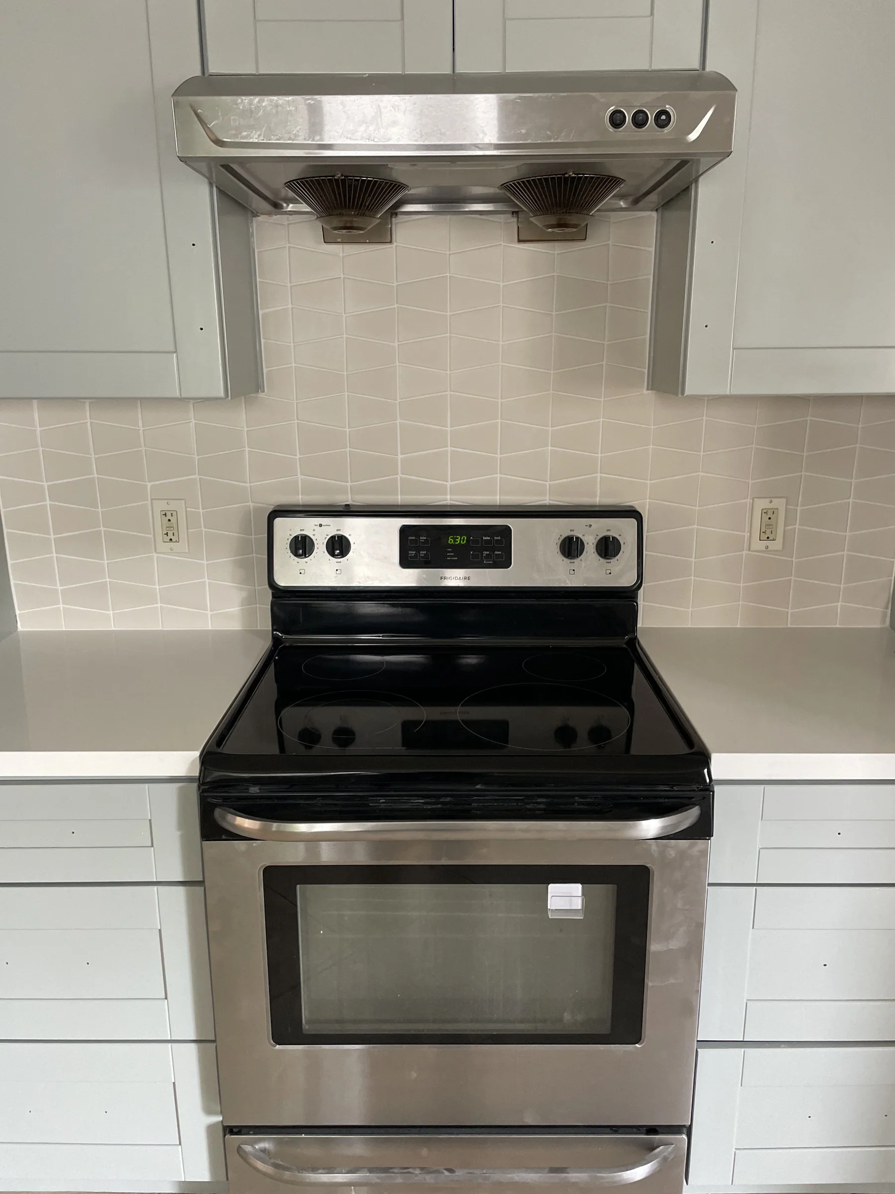 Finished Alameda kitchen range area with white-on-white chevron tile backsplash, white quartz countertop, white shaker cabinets, and a stainless Frigidaire electric range with range hood